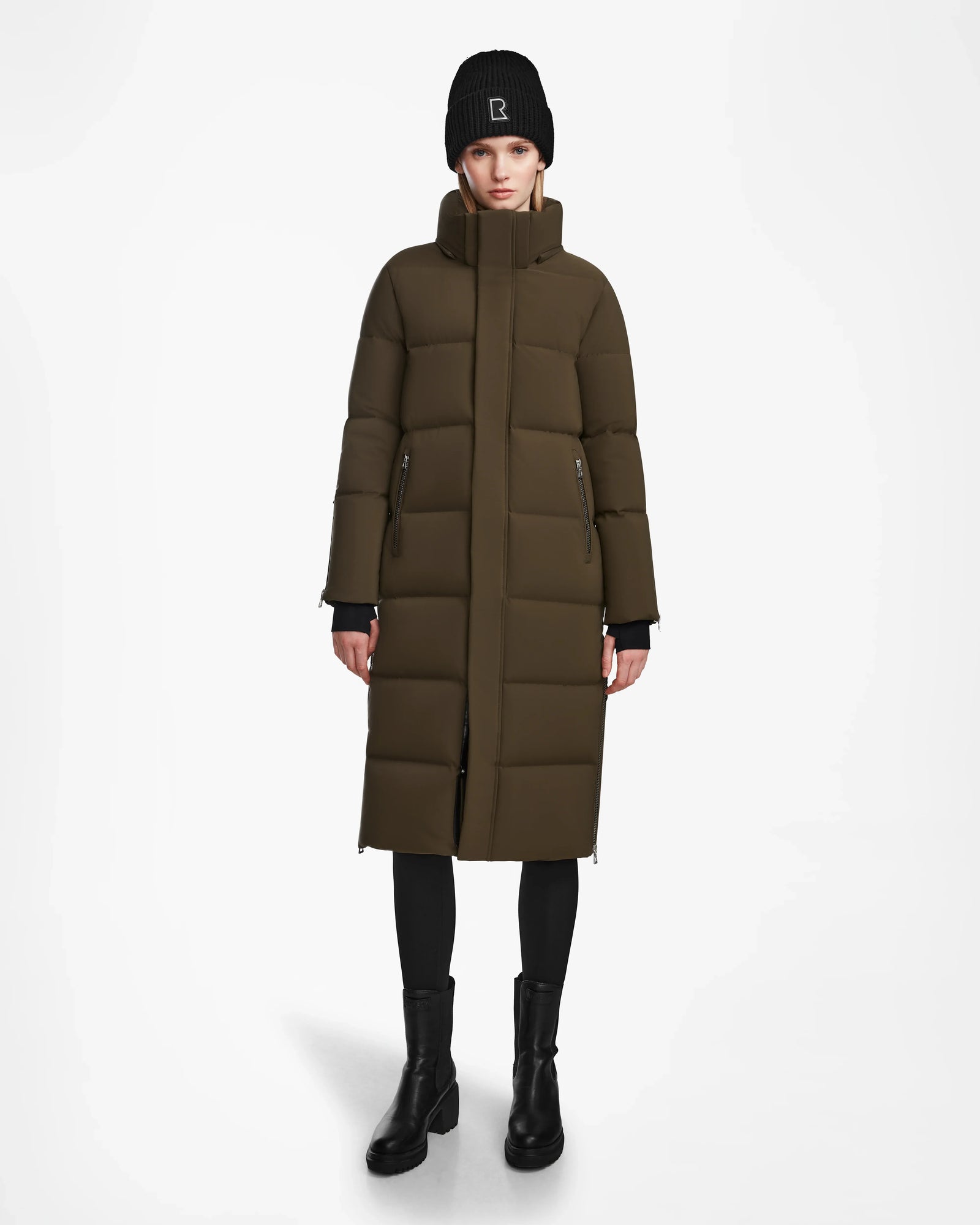 A person wearing a Rudsak MAO M - 8123598 Army womens down puffer coat in olive-green, with weatherproof recycled Stormshell, plus a black knit beanie, black leggings, and boots stands against a plain white background.