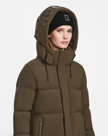 A woman stands against a plain white background wearing the Rudsak MAO M - 8123598 Army olive green winter coat with a hood and a black beanie, both featuring metallic RUB lettering.