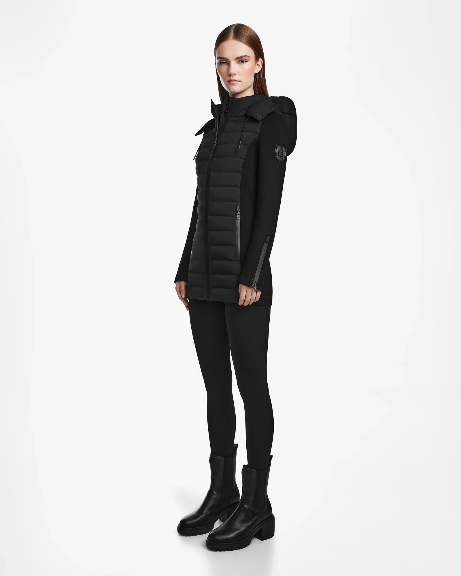 A woman with straight brown hair models the Rudsak MARRY M - L123548 Black womens puffer jacket with removable hood, styled with black leggings and boots, standing against a plain white background.