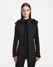 A woman with straight brown hair wears the Rudsak MARRY M - L123548 Black women’s puffer jacket, featuring a removable hood, over a black sweater and leggings against a plain white background.