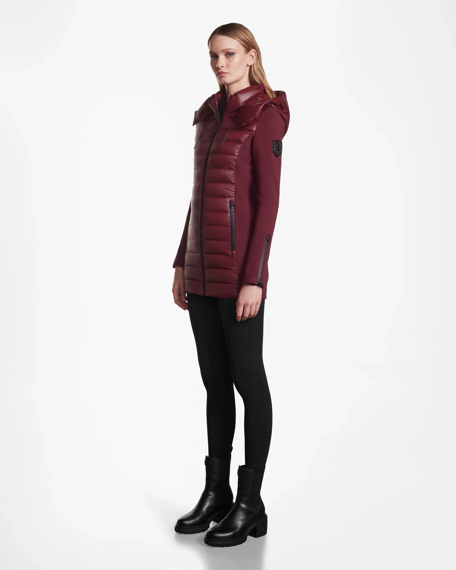 A woman in the Rudsak MARRY - L121003 Wine long puffer vest with hood poses against a white background, paired with black leggings and ankle boots, looking slightly to the side with a neutral expression.