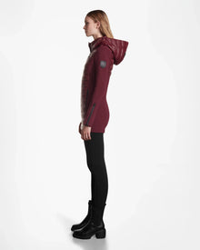 A woman stands in profile against a white background wearing the Rudsak MARRY - L121003 Wine womens puffer coat, paired with black leggings and black ankle boots.