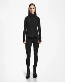 A woman faces forward, wearing the Rudsak MATILDA - L124504 Black vest over a black long-sleeve top, leggings, and boots—an ideal transitional weather look—set against a plain white background.