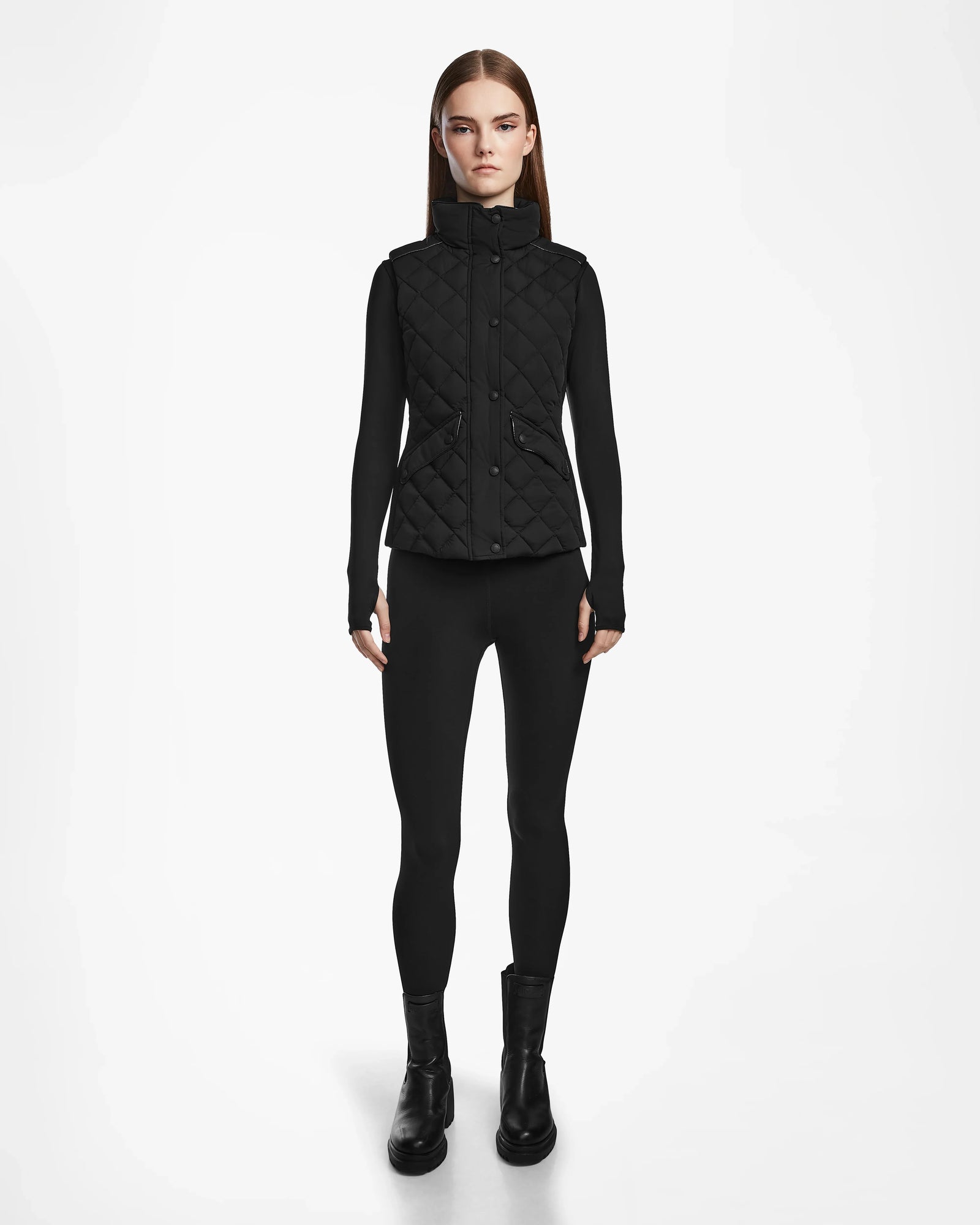 A woman faces forward, wearing the Rudsak MATILDA - L124504 Black vest over a black long-sleeve top, leggings, and boots—an ideal transitional weather look—set against a plain white background.