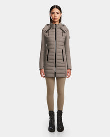A woman faces forward against a white background, wearing the Rudsak MERINA M - 8124030 Cement taupe quilted down jacket with a high collar, tan leggings, and black lace-up boots.