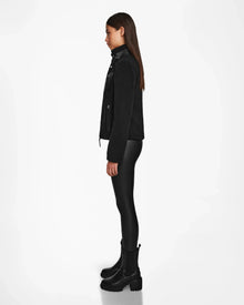 A young woman stands in profile against a white background, wearing the Rudsak MILANA - 8524604 Black zip-up jacket with vegan leather details, fitted black pants, and chunky black boots. Her straight hair is left loose.