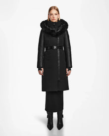 A woman in a Rudsak MISALA - L124512 Black water-resistant down coat with a fur-lined hood and belt stands against a white background, her straight blonde hair and black heeled boots complementing the stylish winter look.