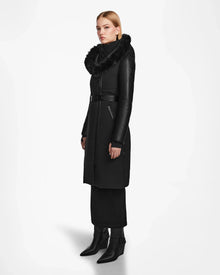 A woman wears the Rudsak MISALA - L124512 Black long down coat with a belted waist and fur-lined collar, styled with a black skirt and pointed boots, standing against a plain white background.
