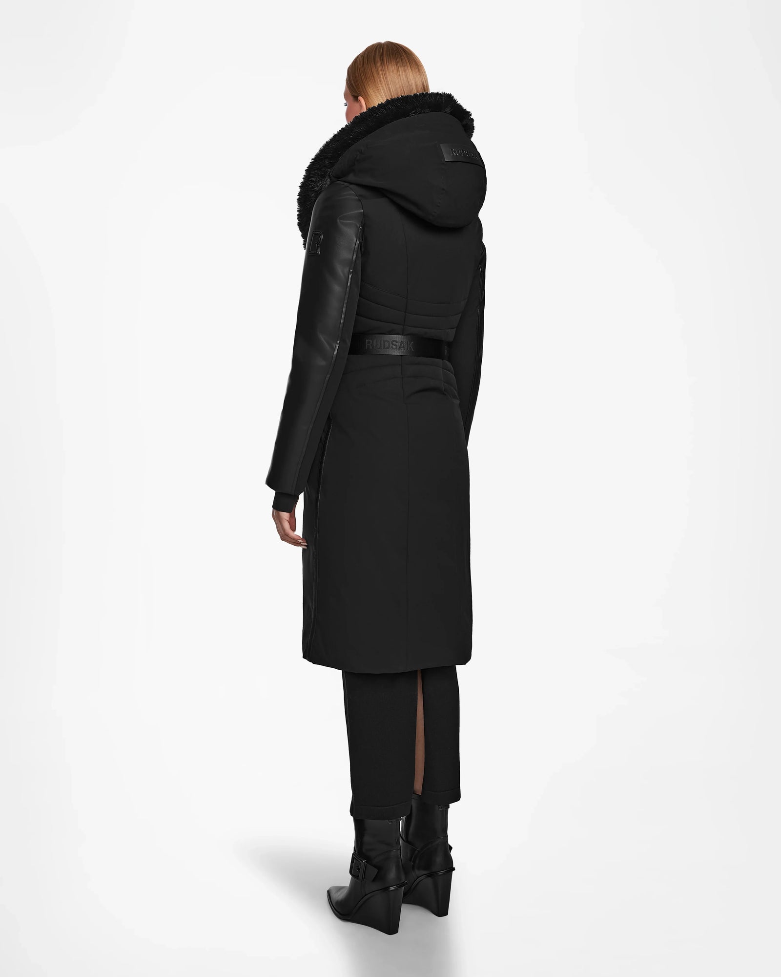 A person with straight, light brown hair is shown from the back wearing the Rudsak MISALA - L124512 Black womens long down coat with a hood, black pants, and black heeled boots against a plain white background.