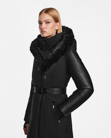 A woman wearing the Rudsak MISALA - L124512 Black long down coat with a belted waist and fur-lined hood poses against a white background, gazing slightly to the side with a neutral expression.