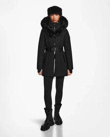 A woman faces forward in the Rudsak MODA M - 8124555 Black down parka with a fur-lined hood, black leggings, lace-up boots, and a matching fur hat. The fitted coat creates a stylish silhouette against a plain white background.
