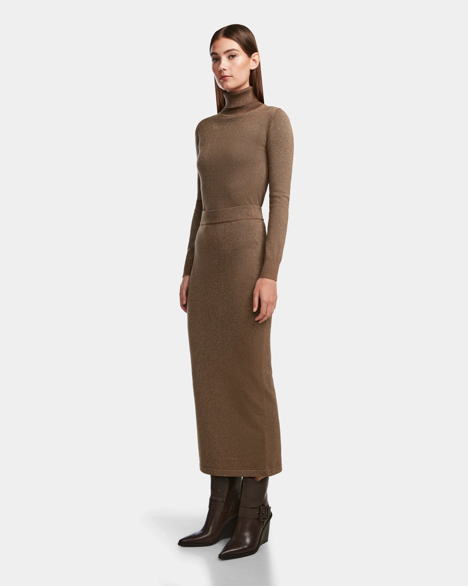 A woman stands sideways in a body-hugging, ankle-length MONICA - 8524516 Antique Bronze knit dress by Rudsak with long sleeves and a turtleneck, paired with dark brown heeled boots, against a plain white background.
