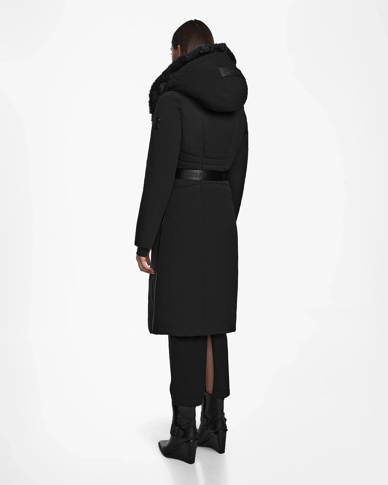 A person faces away, wearing the Rudsak MUSA - L124534 Black women’s long puffer coat with RDS-certified down, fur-trimmed hood, belted waist, black pants, and boots against a plain white background.