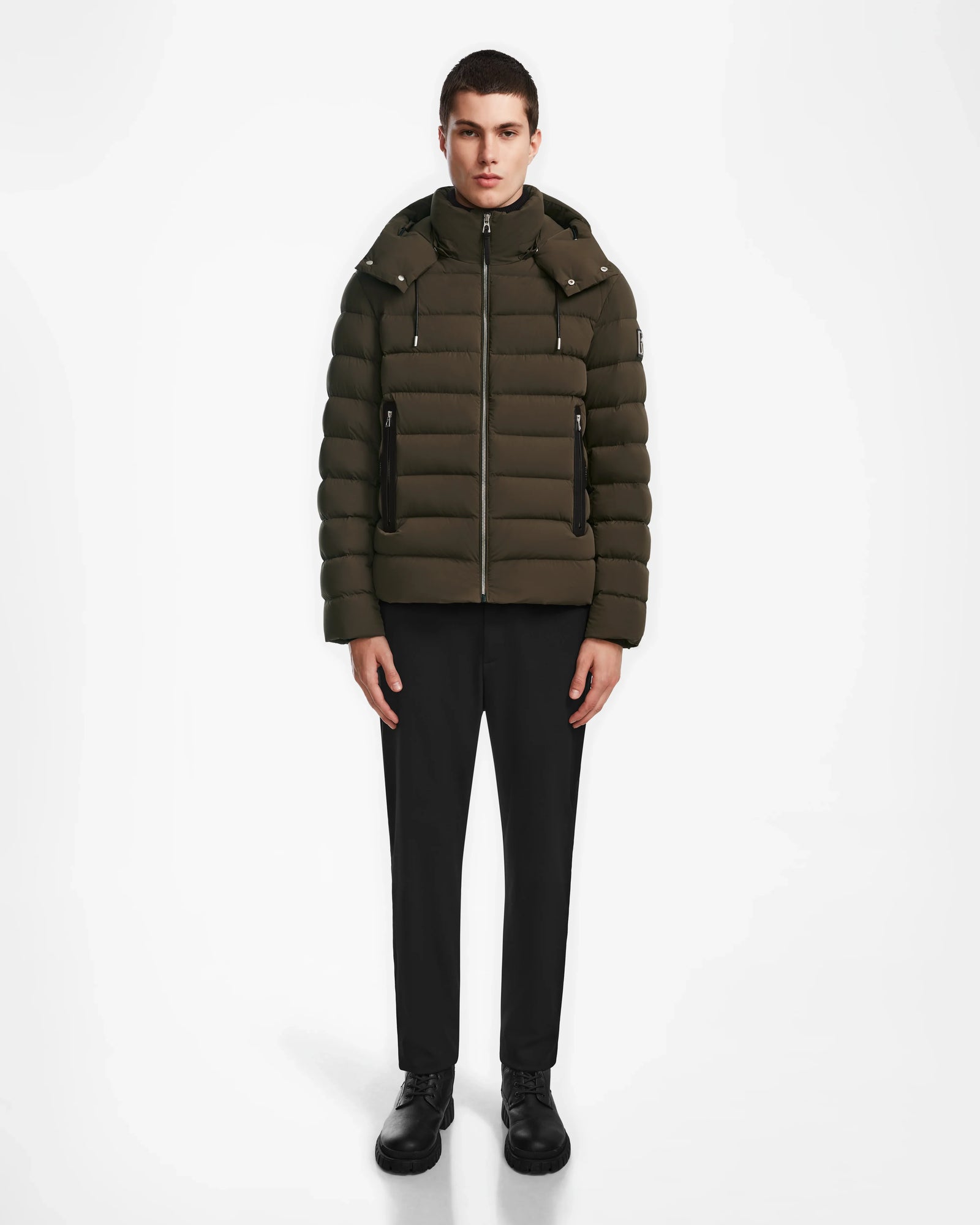 A person faces forward against a plain white background, wearing the Rudsak NAVAN - 6124551 Army mens light down puffer with a hood, paired with black pants and black shoes.