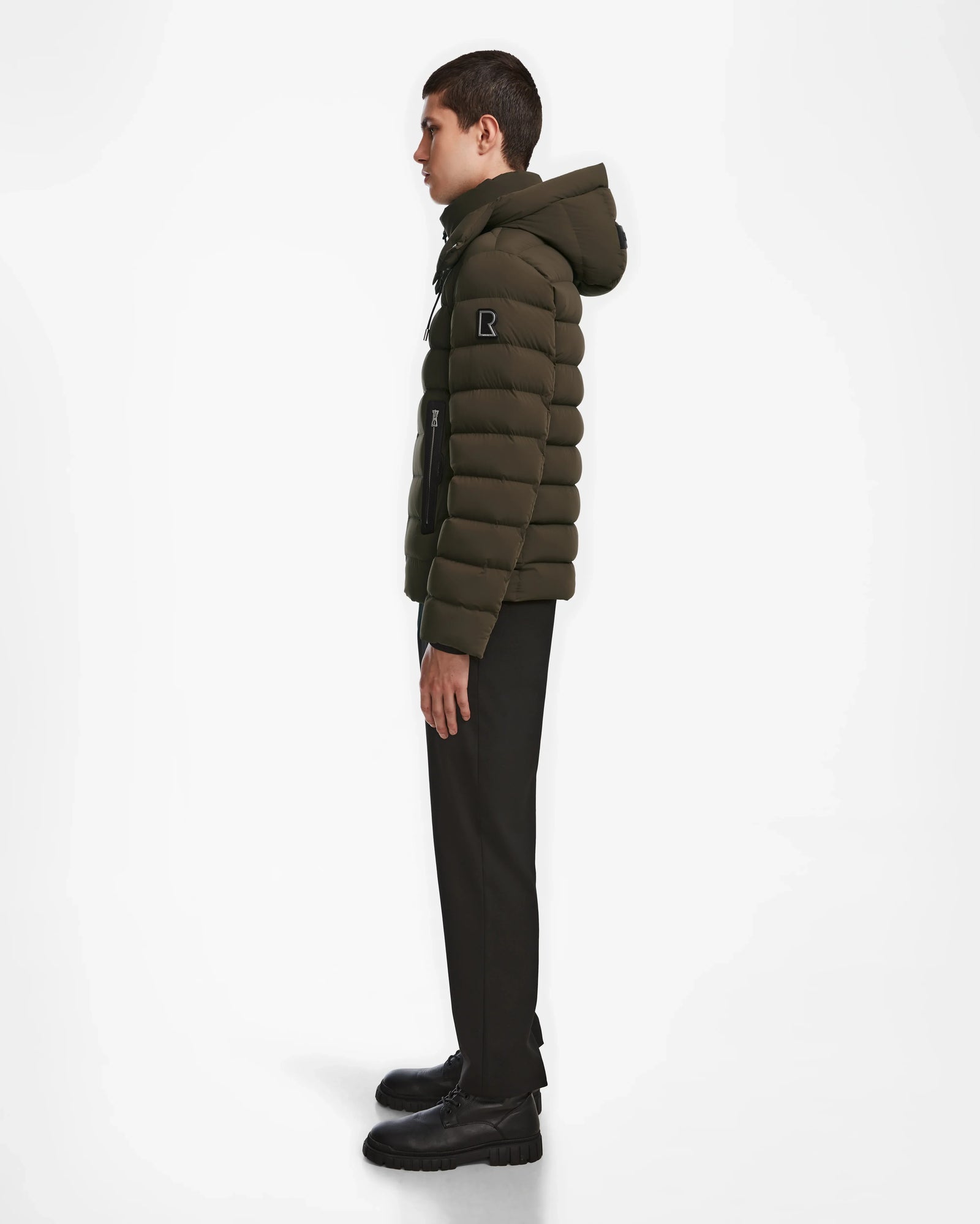 A young man stands in profile against a white background, wearing the Rudsak NAVAN - 6124551 Army mens light down puffer jacket with a hood, along with black pants and black shoes.