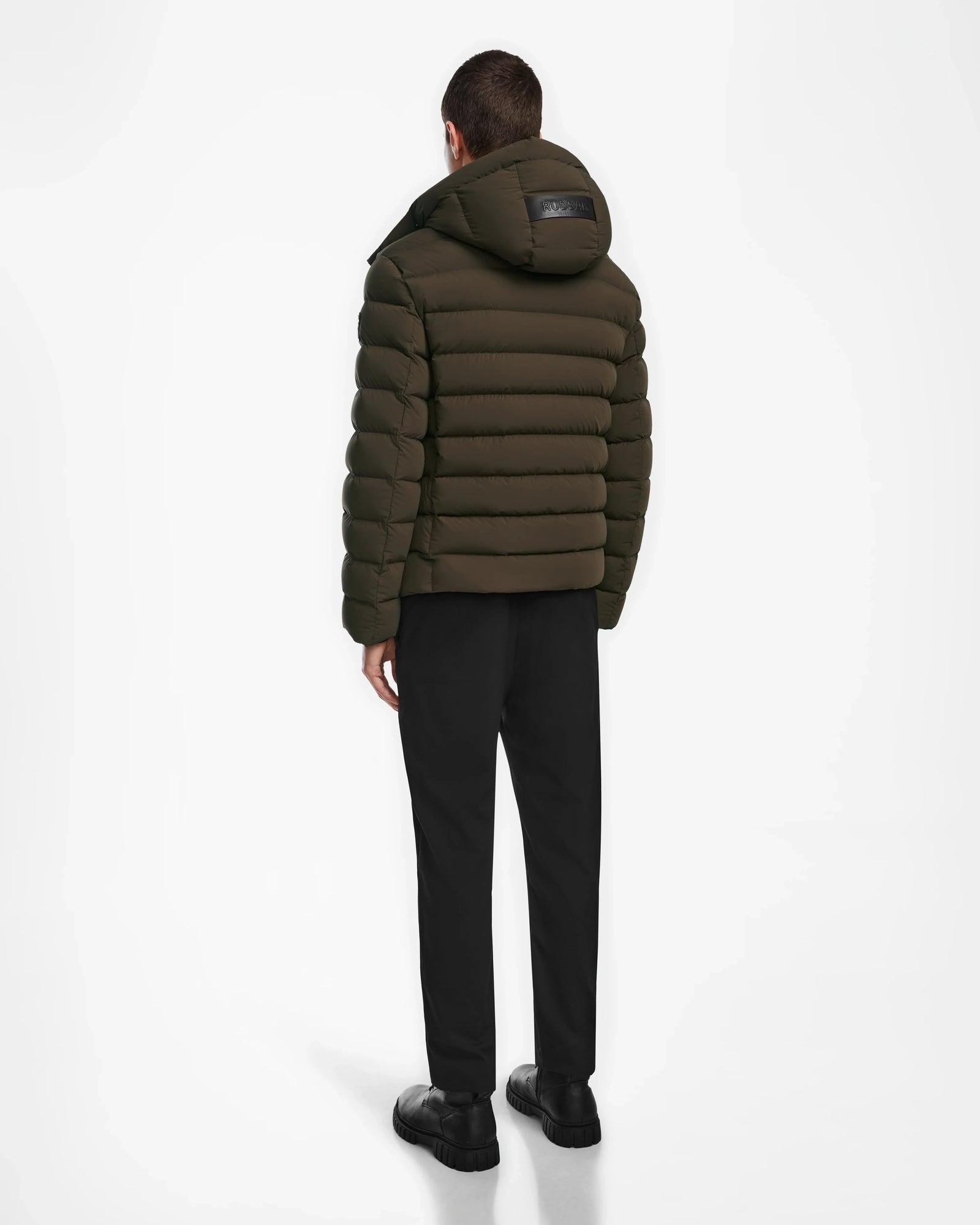 A person stands facing away, wearing the Rudsak NAVAN - 6124551 Army dark olive quilted puffer coat with a hood, paired with black pants and black shoes against a plain white background.
