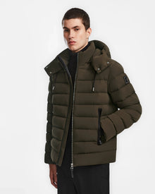 A young man wears the Rudsak NAVAN - 6124551 Army mens quilted down puffer jacket with hood, zipper, and black accents, standing against a plain white background.