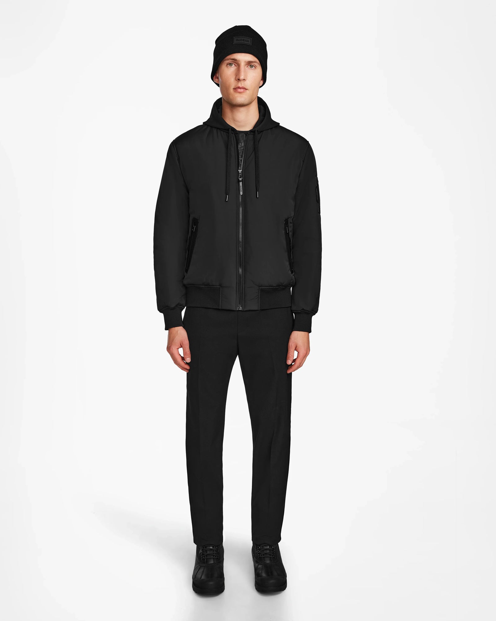 A man stands facing forward against a white background, wearing the Rudsak NEVEN - M124520 Black mens bomber jacket made from recycled materials, paired with a black beanie, black pants, and black shoes. His hands are by his sides and his expression is neutral.