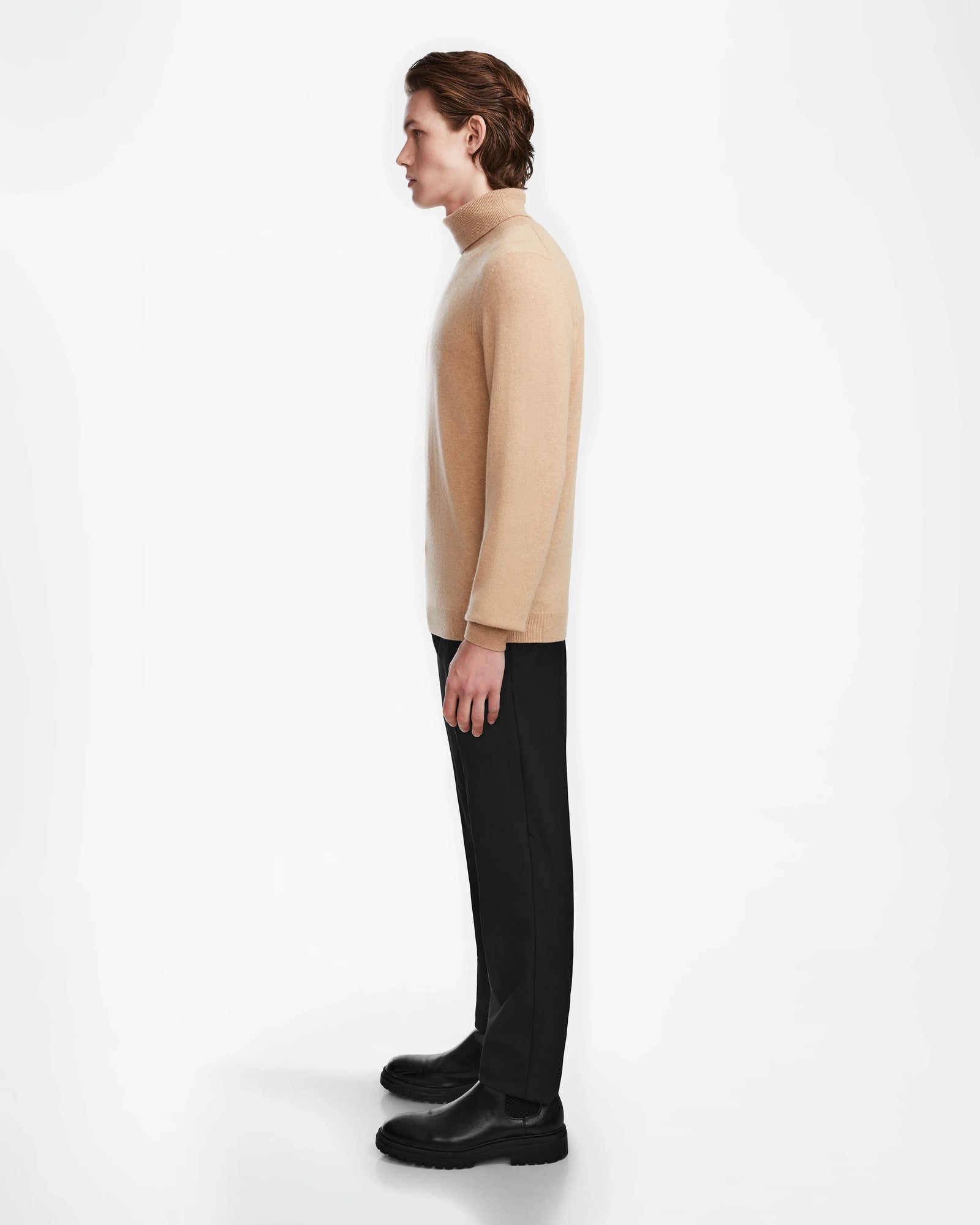 A person with medium-length brown hair stands in profile, wearing the Rudsak NICOLAS - 6524528 Camel men’s turtleneck sweater, black pants, and black shoes, against a plain white background.