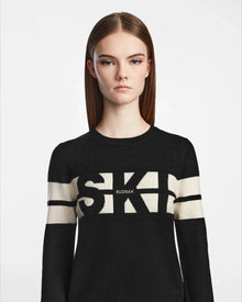 A woman with straight brown hair and a neutral expression wears the Rudsak ODESSA - 8523540 Black soft-knit sweater, featuring bold white SKI and RUDSAK lettering across the chest and arms. She stands against a plain white background.
