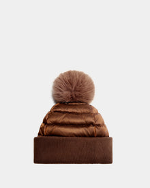 The Rudsak OLYA - 8422504 Antique Bronze is a women’s winter puffer beanie in brown, featuring a wide knit cuff and a large fur pompom, shown against a plain light background.
