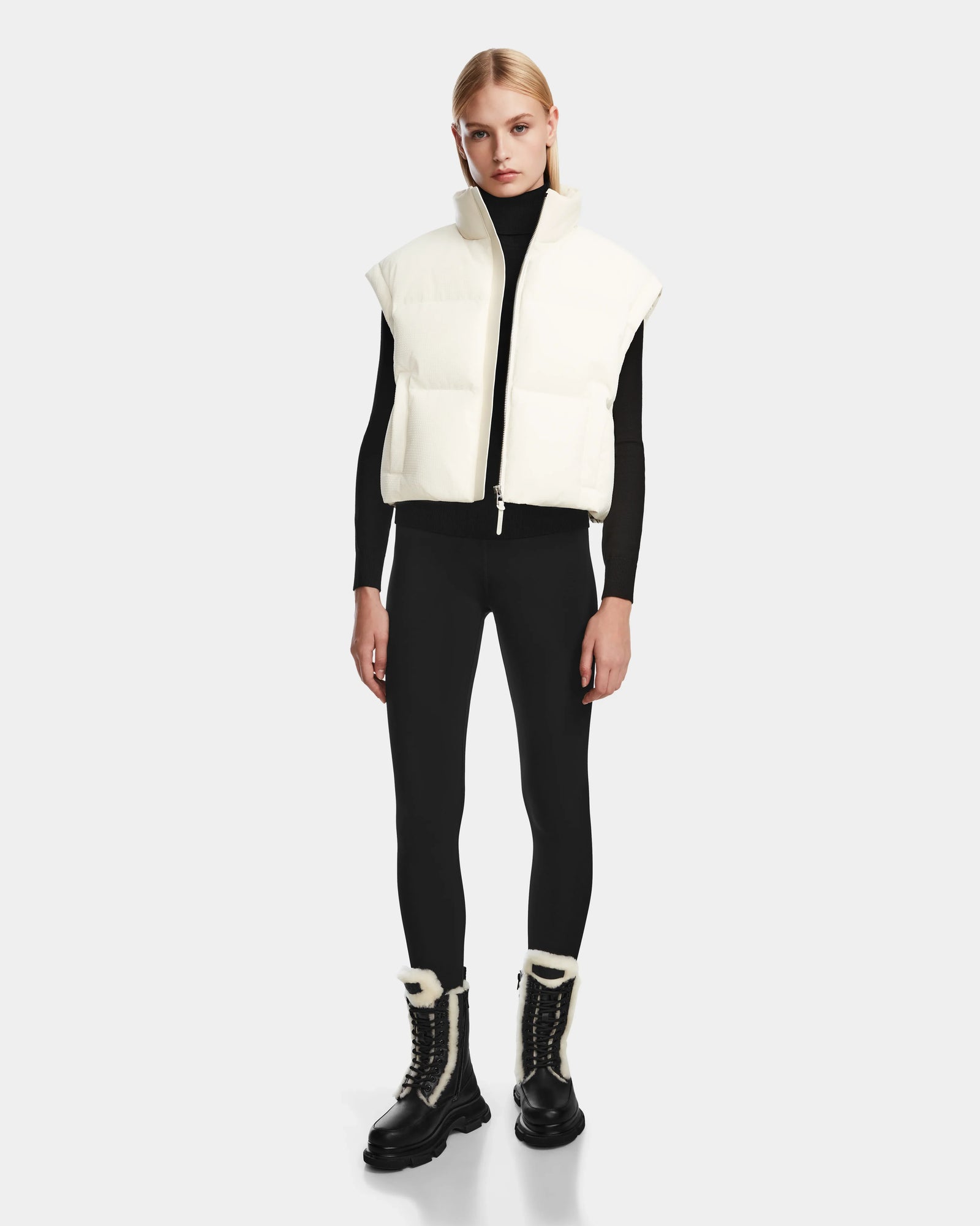 A woman faces forward wearing the Rudsak OPRAH - 8124708 Iceland womens cropped down puffer vest over a black turtleneck, leggings, and black lace-up boots with white trim, against a plain light gray background.