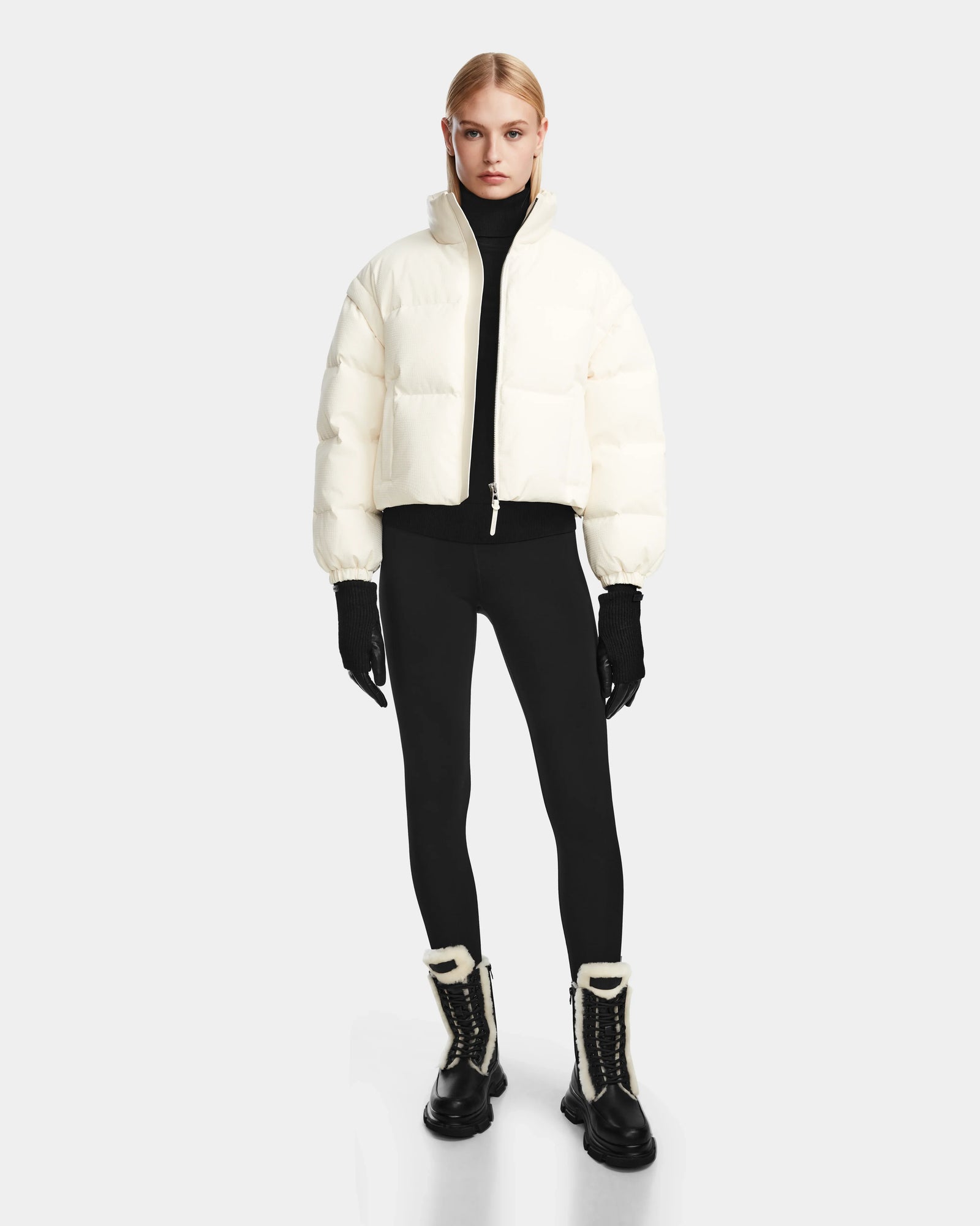 A person stands facing forward wearing the Rudsak OPRAH - 8124708 Iceland womens cropped down puffer jacket with removable sleeves and RDS certified ethical down, paired with a black turtleneck, leggings, gloves, lace-up boots with white fur trim.