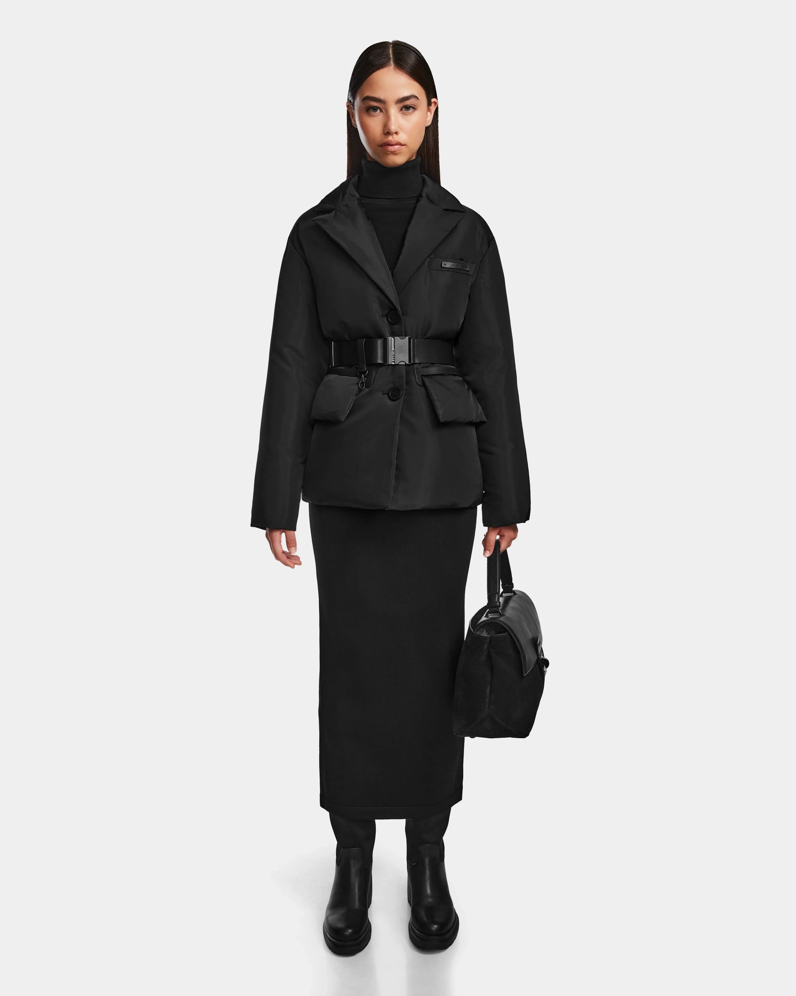 A woman with straight dark hair and a neutral expression stands against a white background, wearing the Rudsak PAOLA - 8124547 Black womens down blazer over a black turtleneck dress, black boots, and holding a black handbag.
