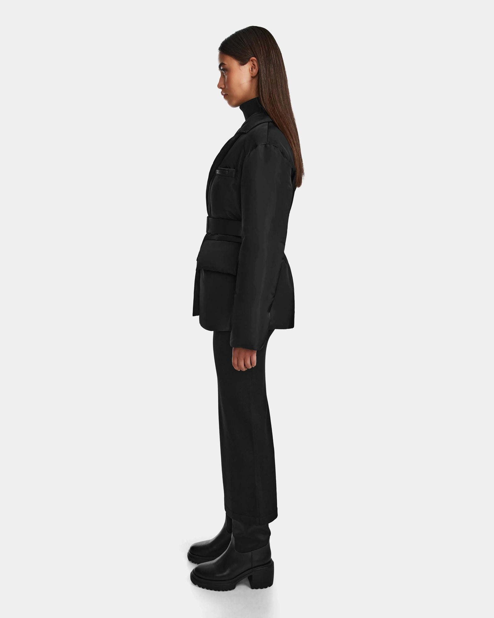 A woman with long brown hair stands in profile facing left, wearing the Rudsak PAOLA - 8124547 Black weather-resistant tailored blazer, paired with black pants, a turtleneck, and chunky-heeled boots against a plain light background.