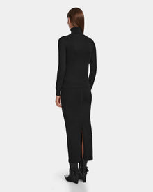A woman with straight brown hair faces away, wearing the RALDI - 8524605 Black by Rudsak—a fitted metallic thread turtleneck—paired with a long black skirt with a back slit and black heeled boots, against a plain white background.