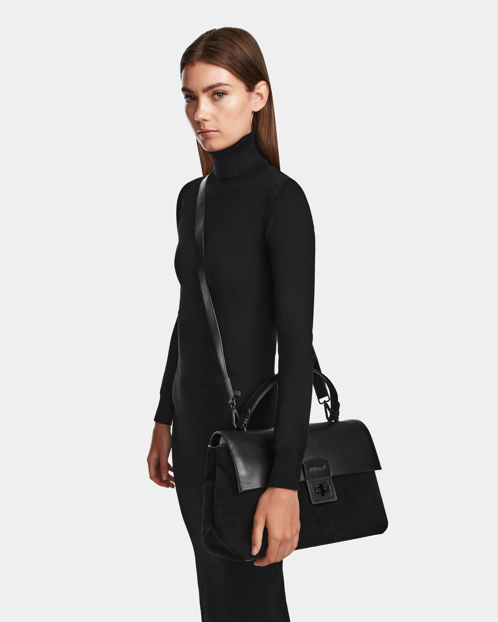 A woman models the RALDI - 8524605 Black fitted turtleneck dress by Rudsak, holding a large black handbag with buckle and shoulder strap against a plain light background.