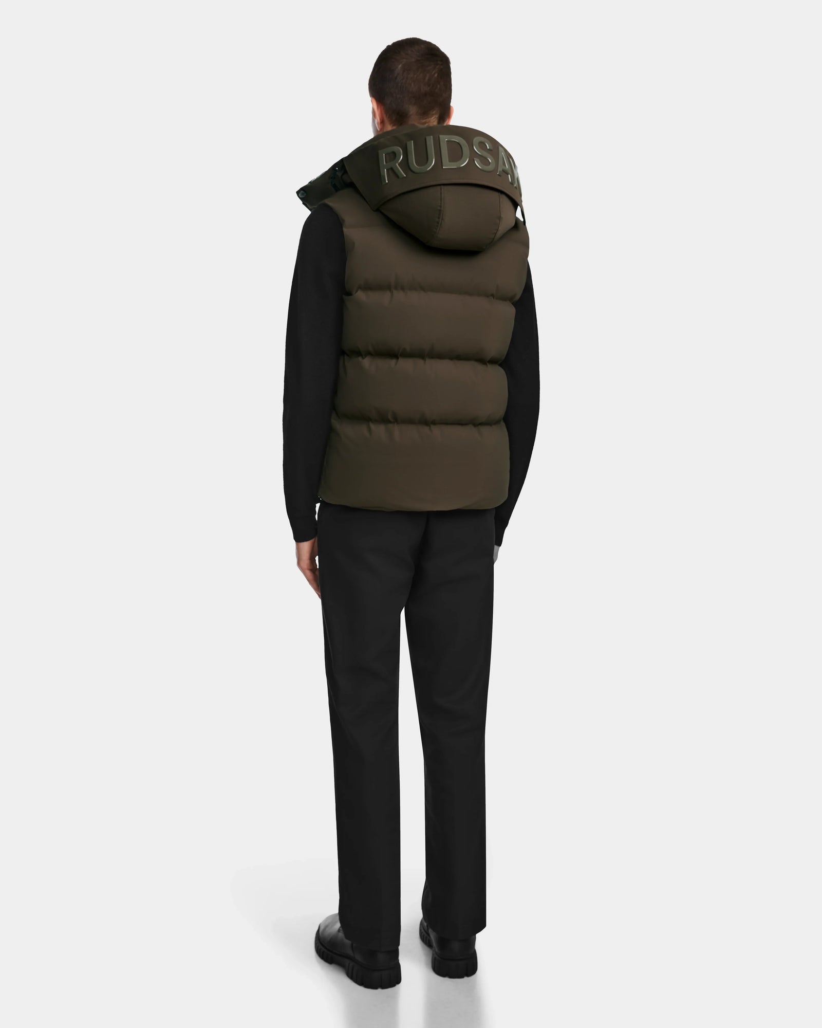 A man faces away in black pants, shoes, and a black long-sleeve shirt layered with the Rudsak REN M - 9123512 Army M olive weather-resistant puffer vest with RDS-certified down. RUDSAK is printed on the hood. Background is plain white.