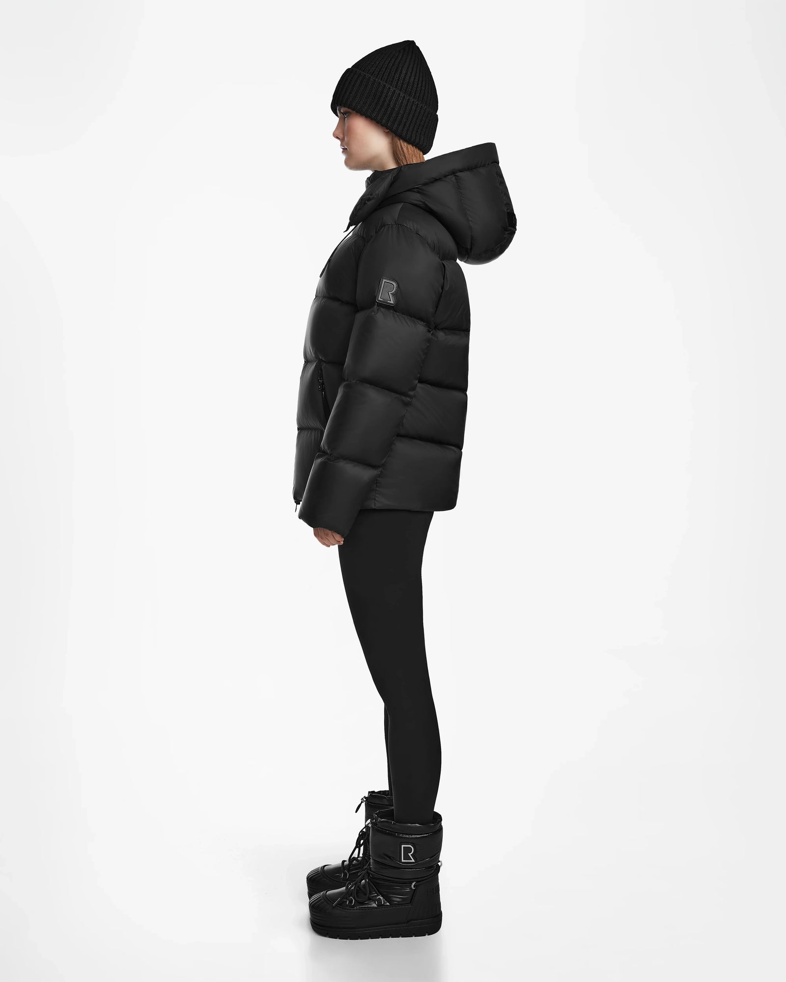 A person in profile stands against a white background, wearing the Rudsak RITA M - L124537 Black weather-resistant puffer jacket with a hood, paired with a black beanie, black leggings, and black winter boots.