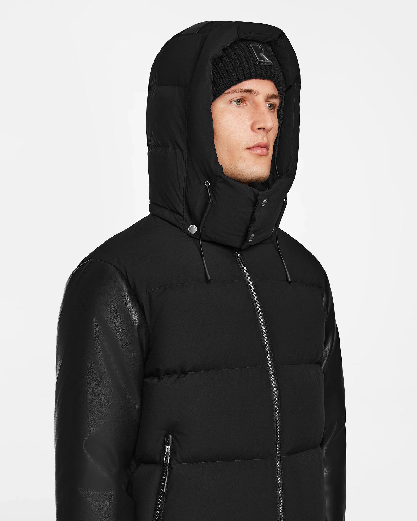 A person wearing the Rudsak RONNY M - M124513 Black puffer jacket with a hood and a black knit beanie, looking slightly to the side against a plain white background.