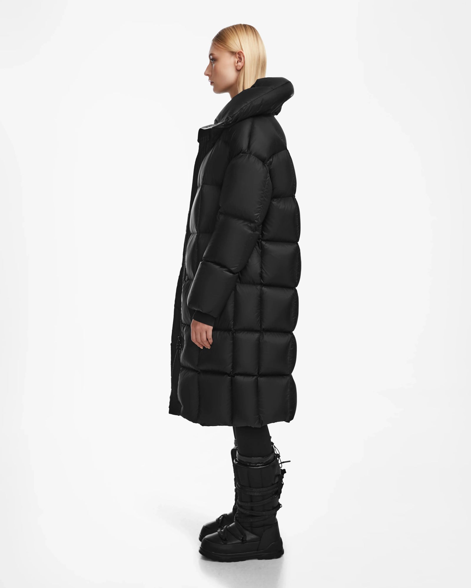 A person with straight blonde hair, wearing the Rudsak SASH - 8124536 Black womens long down puffer coat, stands in profile against a plain white background.