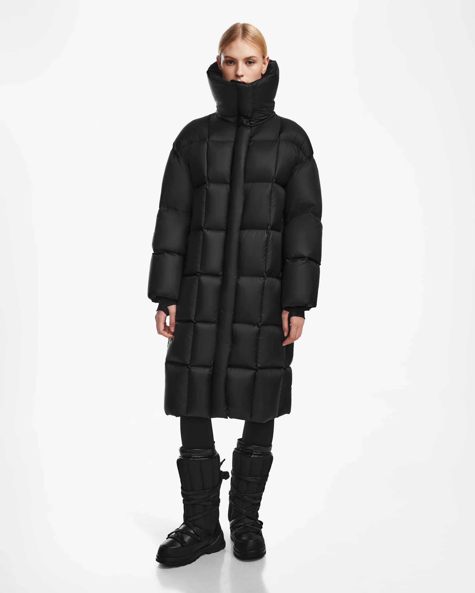 A person stands facing forward, wearing the Rudsak SASH - 8124536 Black women’s long down puffer coat with a high collar, and black winter boots. The coat is filled with RDS-certified down. The background is plain white.