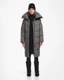 A woman models the Rudsak SASH - 8124536 Light Grey womens long down puffer coat, styled with a black beanie, pants, and winter boots. She stands against a plain white background, facing forward with a neutral expression.