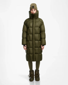 A woman wearing the Rudsak SASH - 8124536 Loden Green long down puffer coat, with a beanie, leggings, and boots, stands facing forward against a plain white background.