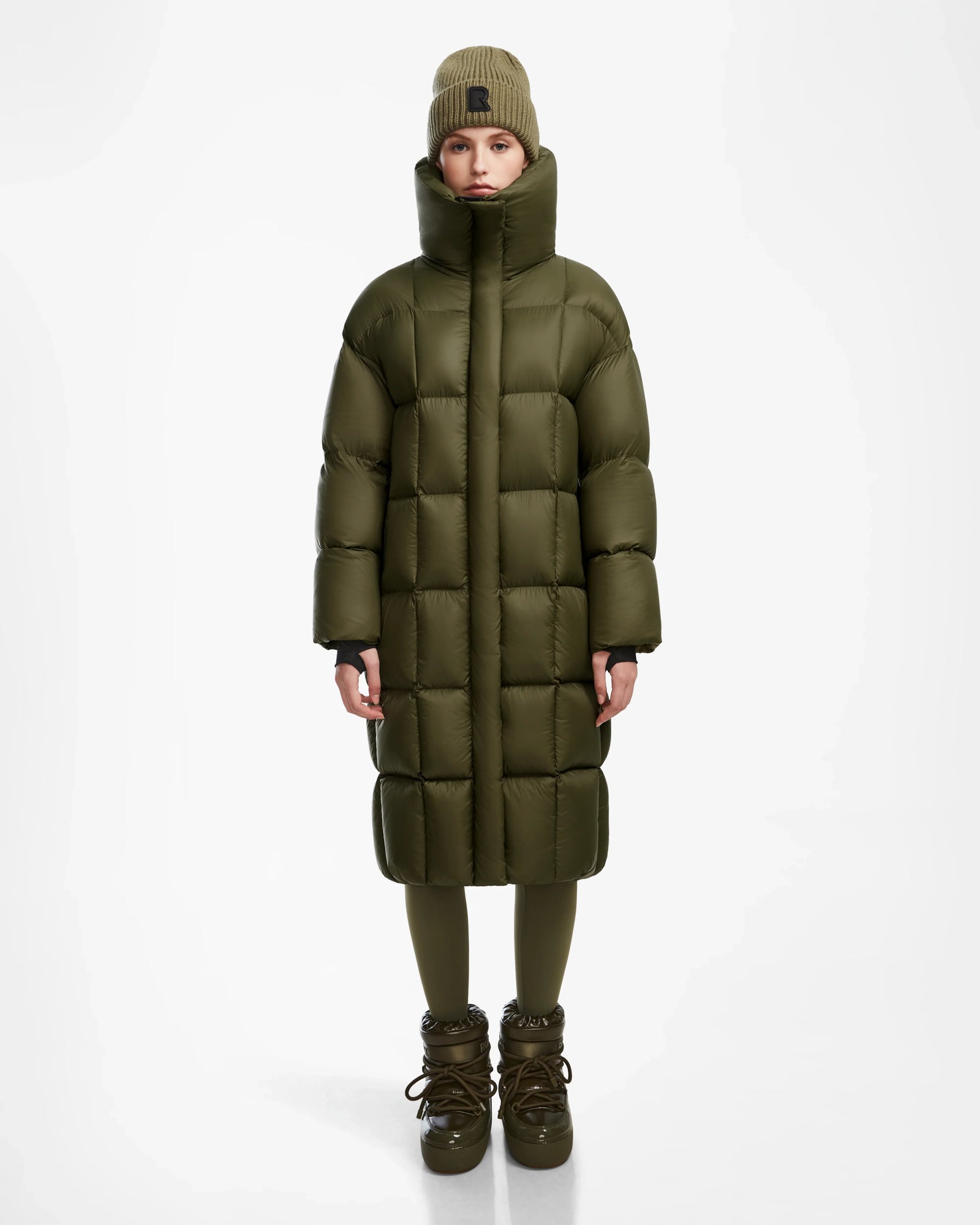 A woman wearing the Rudsak SASH - 8124536 Loden Green long down puffer coat, with a beanie, leggings, and boots, stands facing forward against a plain white background.