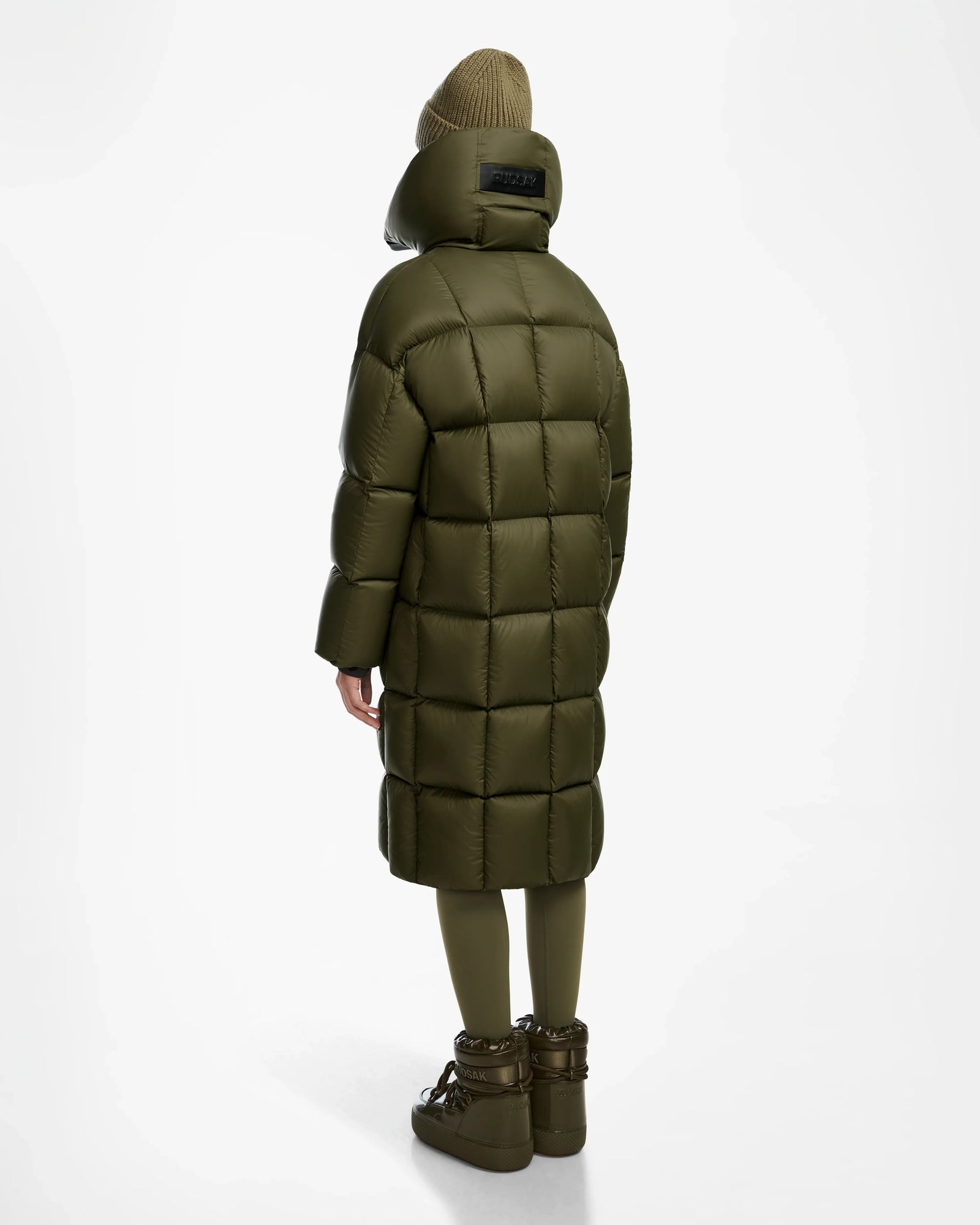 A person stands facing away, wearing the Rudsak SASH - 8124536 Loden Green women’s long down puffer coat with RDS-certified down, paired with matching leggings, chunky boots, and a brown knit beanie against a white background.