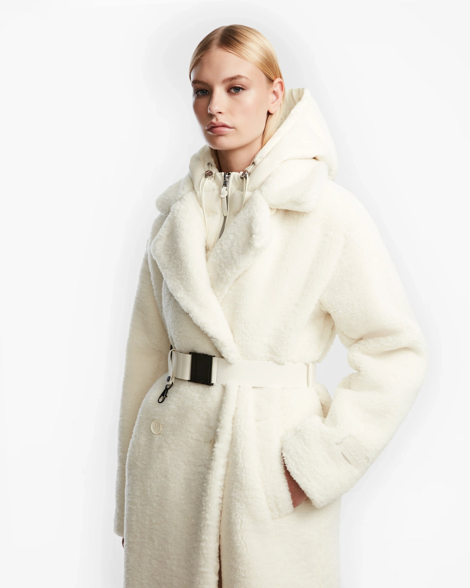 A woman with blonde hair models the Rudsak TINA - 8123578 Iceland: a white belted faux fur trench coat with a large collar and removable hooded bib. She stands against a plain white background, one hand in her pocket.