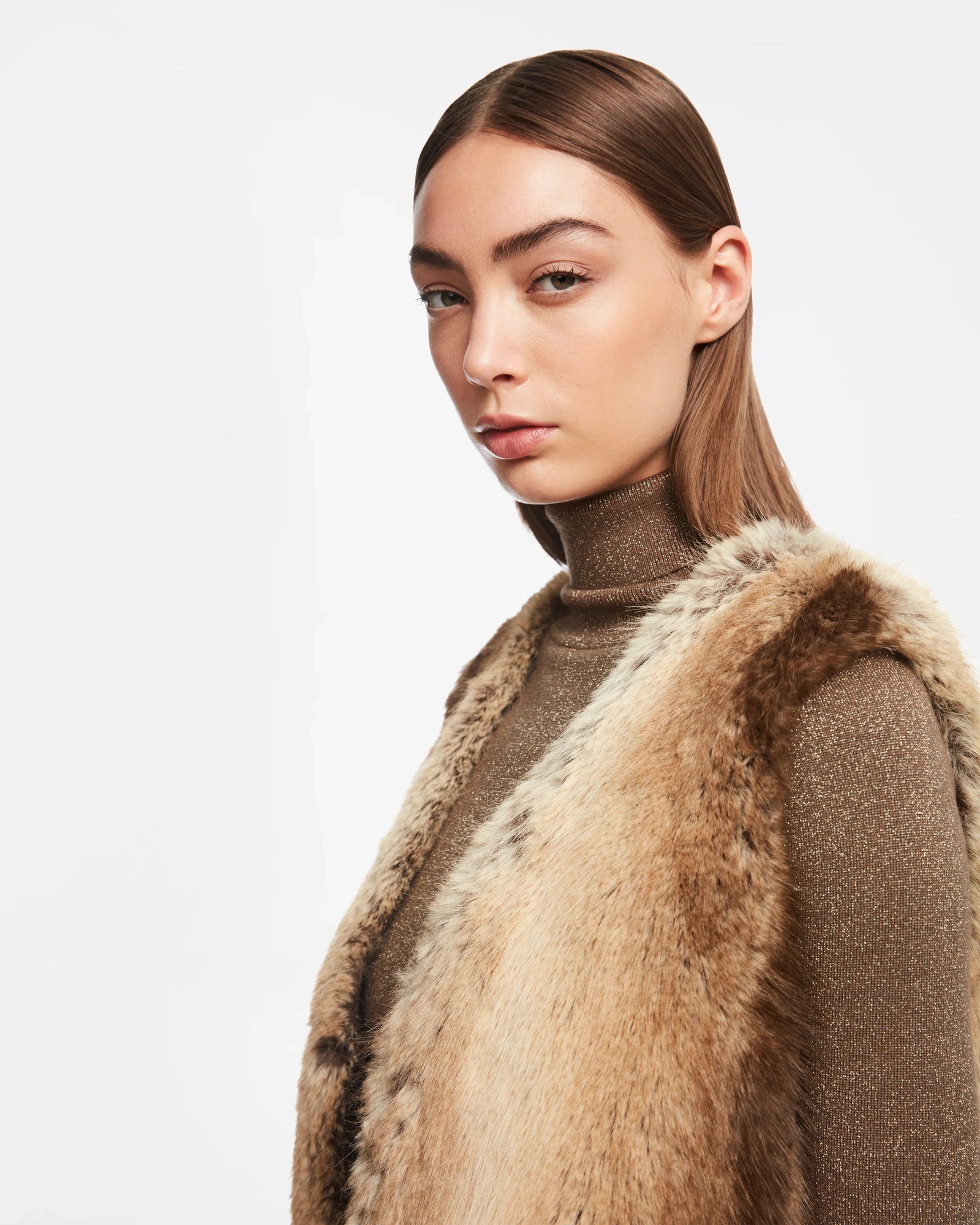 A woman with straight brown hair wears the Rudsak YOLANDA - 8124790 Mocha womens faux fur vest, cinched with a vegan leather belt and paired with a brown turtleneck, staring seriously at the camera against a plain white background.