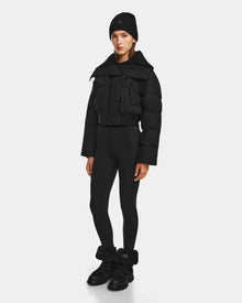 A person stands facing forward, wearing Rudsaks YVETTE - 8124710 Black crop down puffer jacket, paired with a black knit beanie, black leggings, and black shoes with fuzzy ankle cuffs, highlighting chic winter style against a plain white background.
