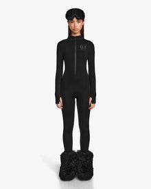A woman faces forward in a fitted Rudsak ZADIE - 8524609 Black long-sleeve bodysuit with a front zipper, black fuzzy slippers, and a black sleep mask on her head, standing against a plain white background.