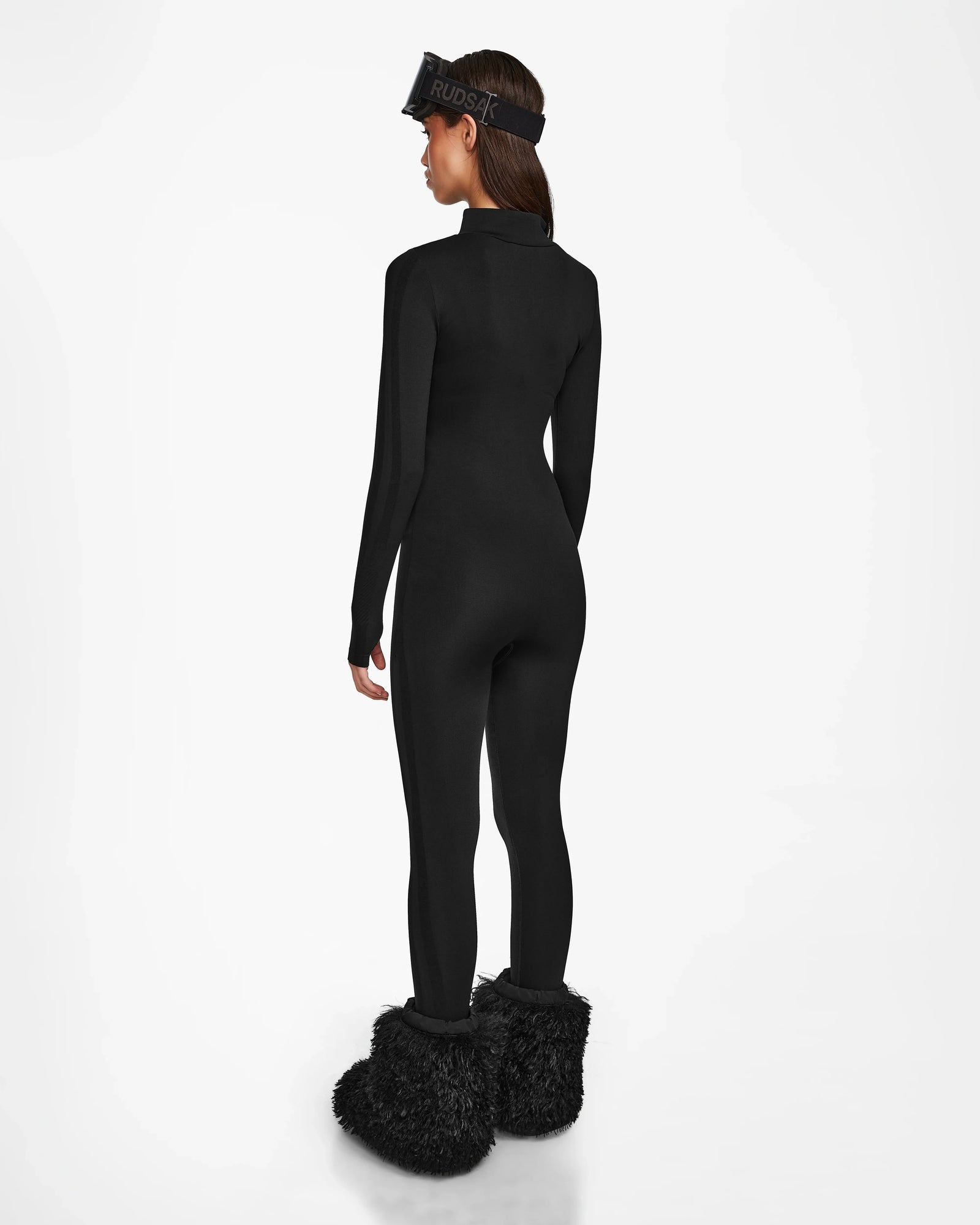 A woman stands facing away against a white background, wearing the ZADIE - 8524609 Black bodysuit and black furry boots, accessorized with a Rudsak-branded black visor.