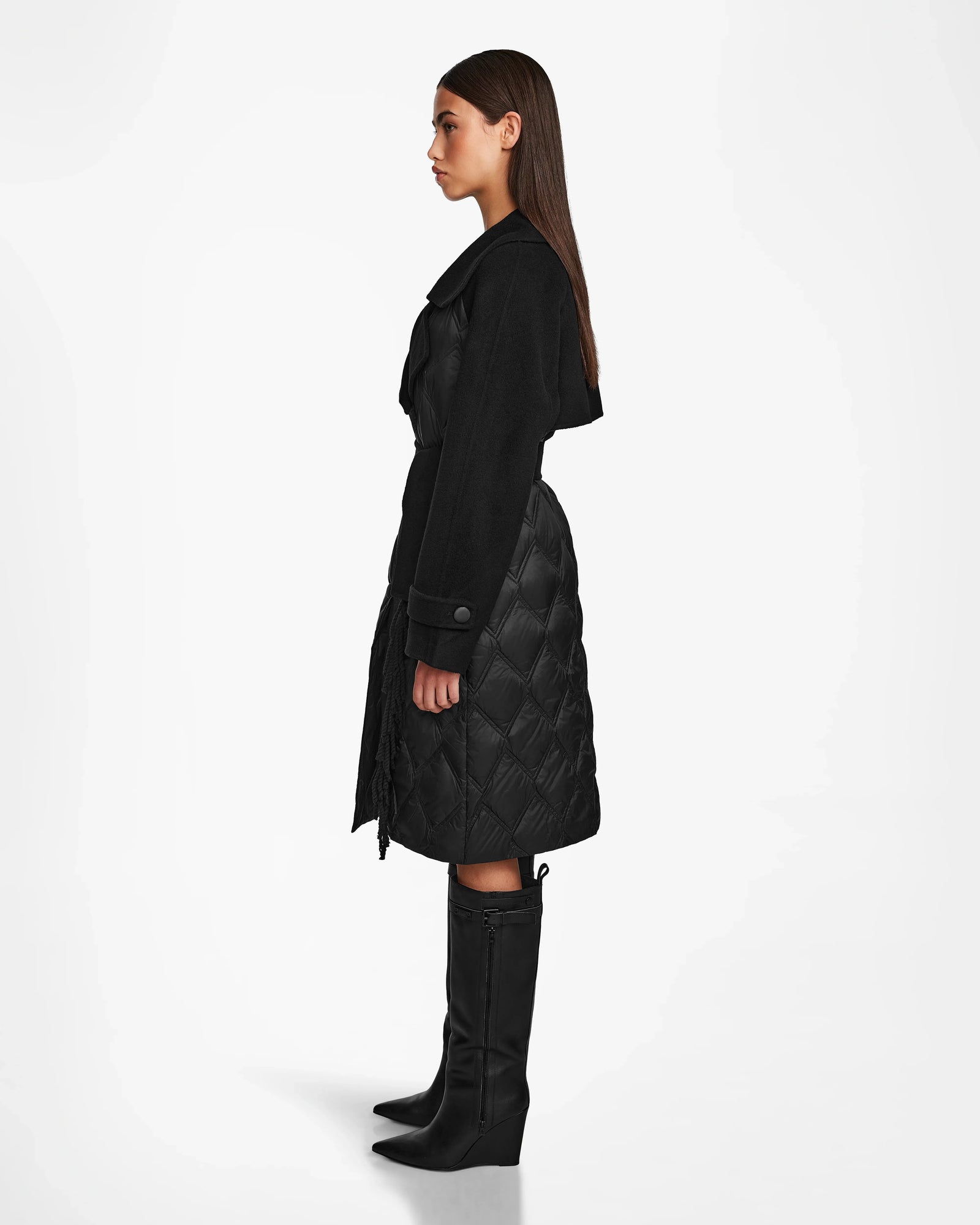A woman in profile stands against a white background wearing the Rudsak ZOE - 8124711 Black long quilted down trench coat and black knee-high boots. Her straight brown hair is down, and her arms rest at her sides.