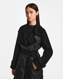 A woman with straight, dark hair models the Rudsak ZOE - 8124711 Black quilted trench coat, featuring a wide collar, button details, and belted waist, standing against a plain white background.
