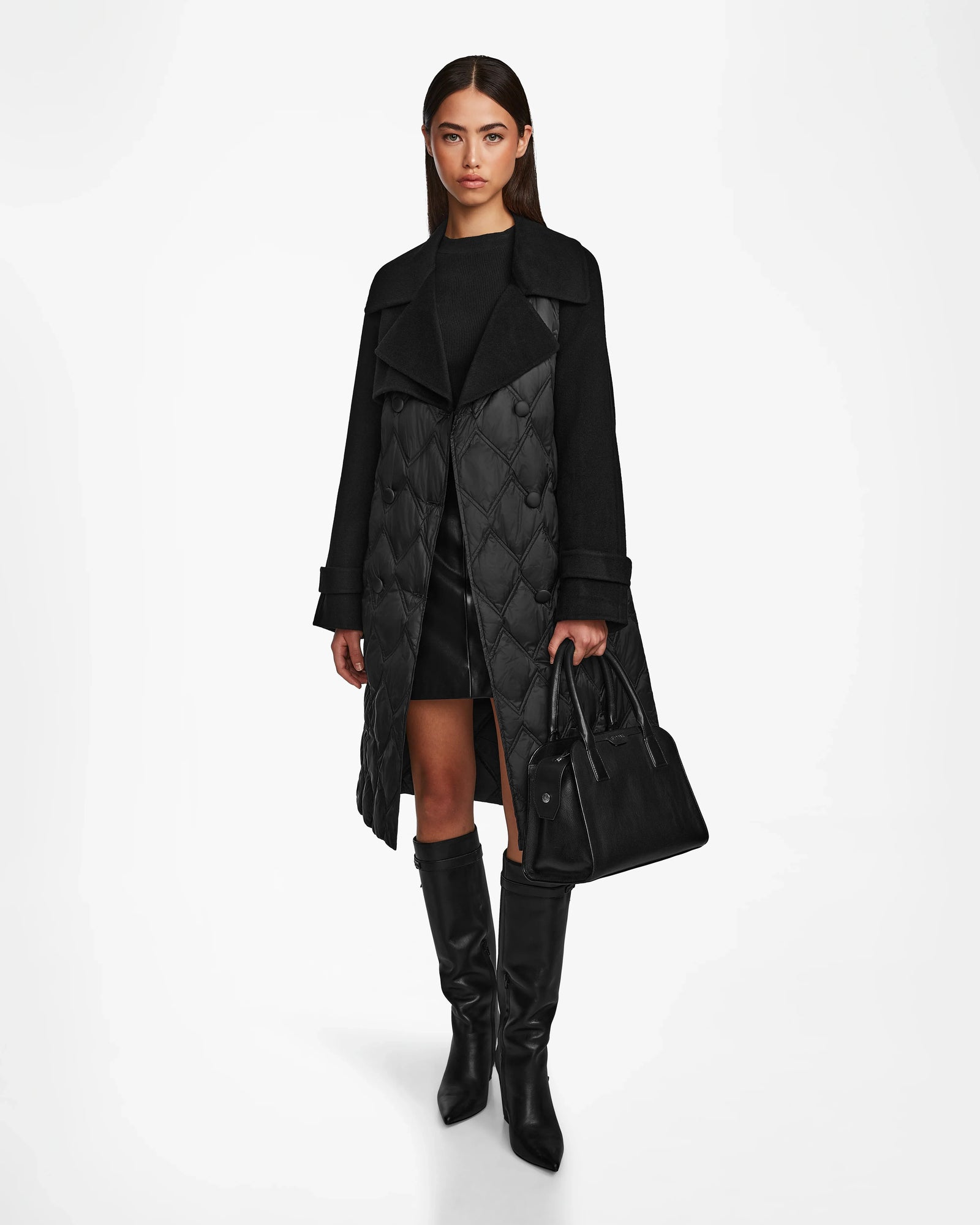 A woman faces forward against a plain white background, wearing the Rudsak ZOE - 8124711 Black womens trench coat with a black dress, knee-high boots, and holding a large black handbag.