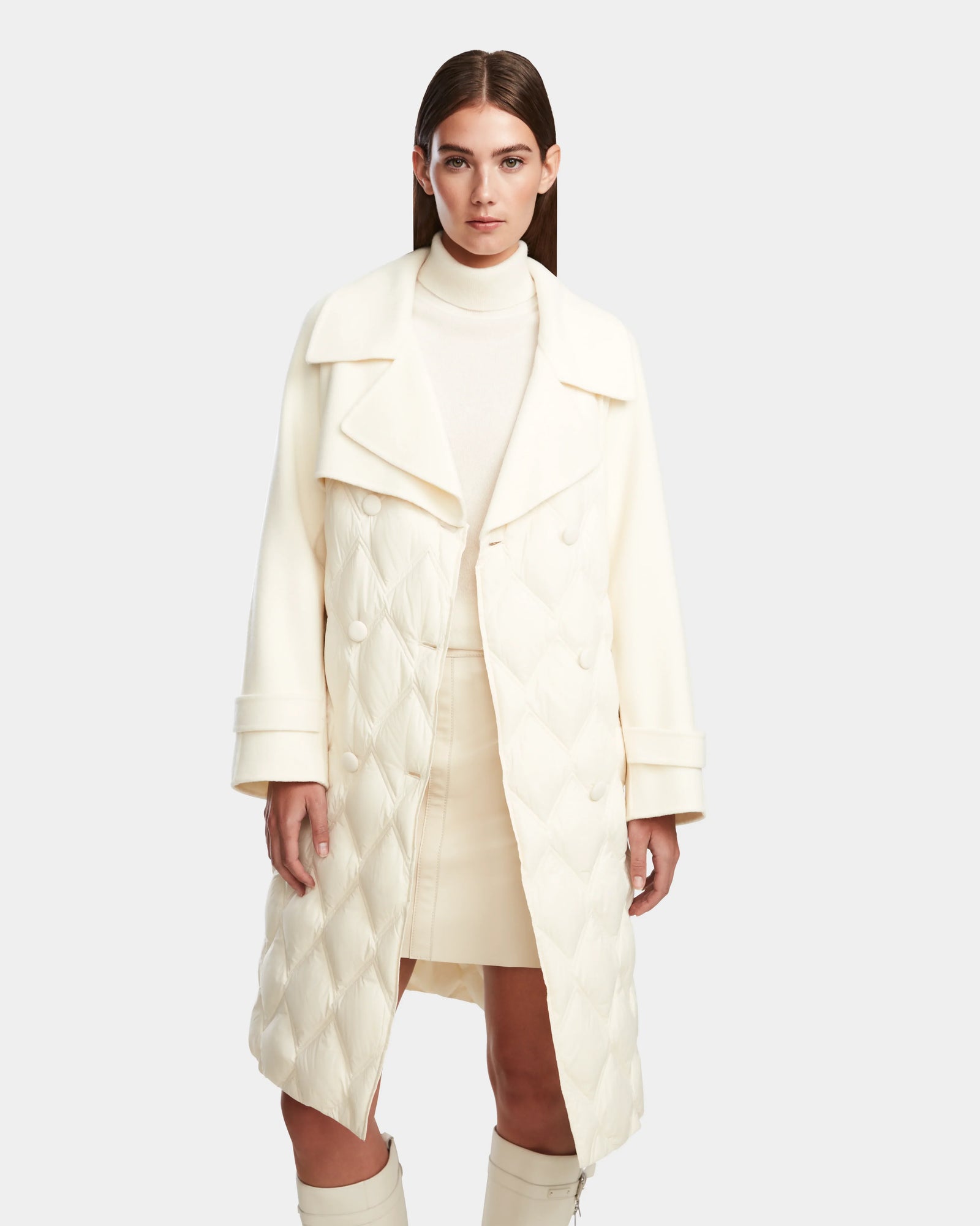 A woman in the Rudsak ZOE - 8124711 Iceland cream quilted down trench with large lapels, layered over a matching turtleneck and skirt, stands against a plain light gray background.