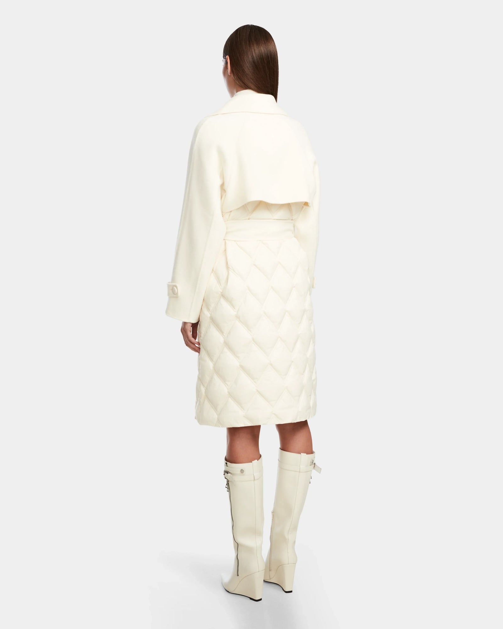 A woman faces away wearing the Rudsak ZOE - 8124711 Iceland, a cream quilted down trench with long sleeves and cape detail, paired with matching knee-high boots.